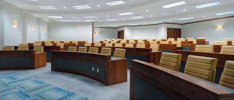 A conference room with rows of wooden desks and beige chairs, equipped with overhead lighting and a patterned carpet.