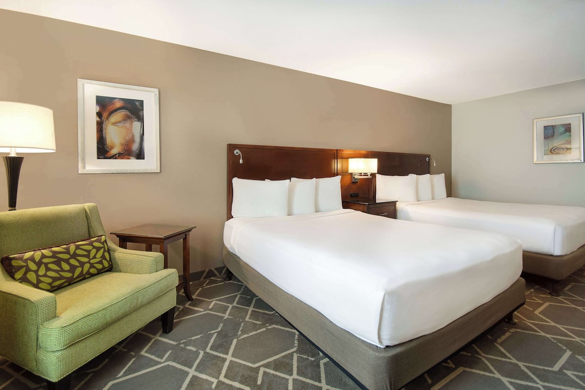 A hotel room with two beds, a green armchair, a lamp, and artwork on the walls.