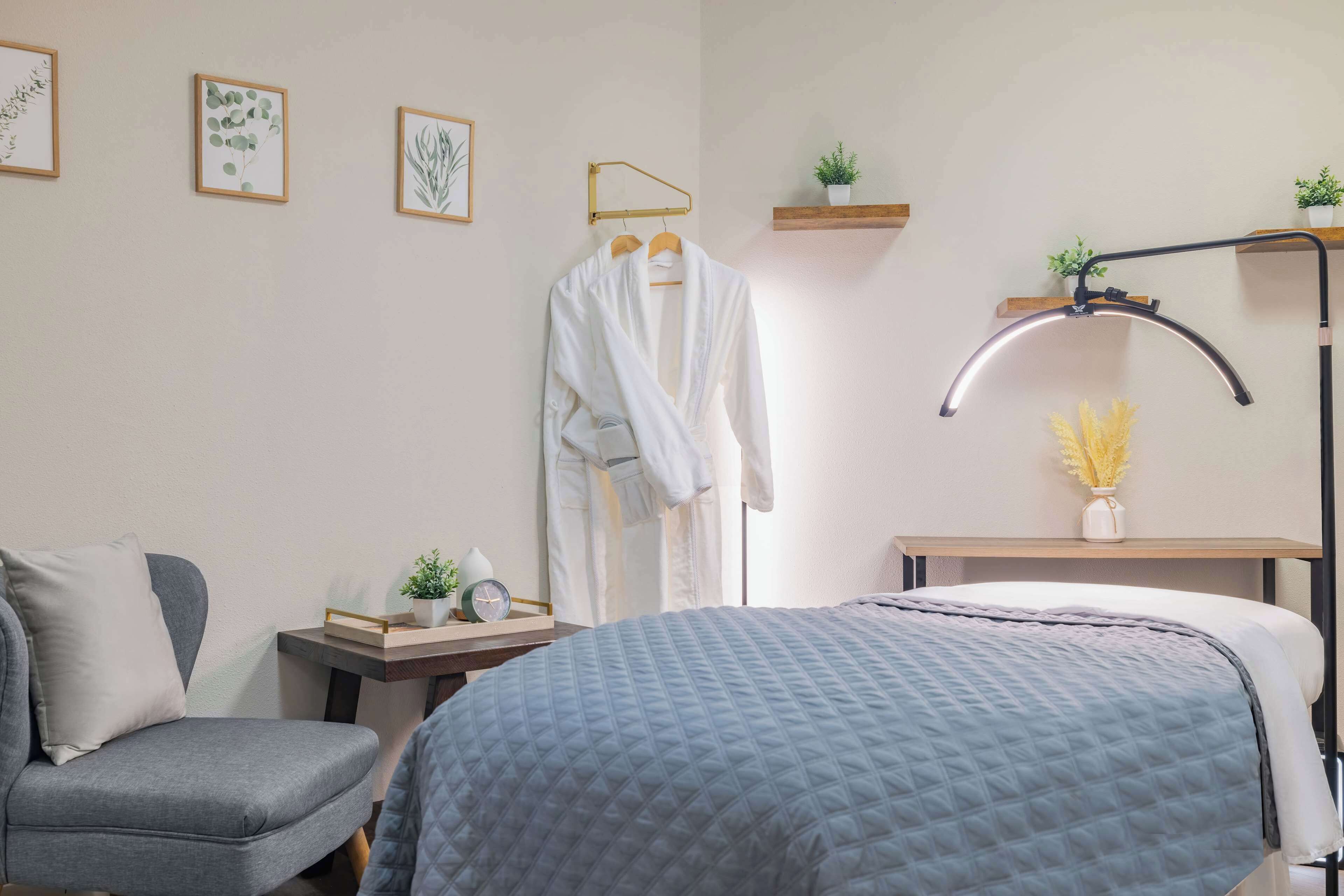 Een keurig opgemaakte slaapkamer met een bed, een stoel, een badjas hangend aan een kapstok, een nachtkastje met een lamp en wandplanken met planten.