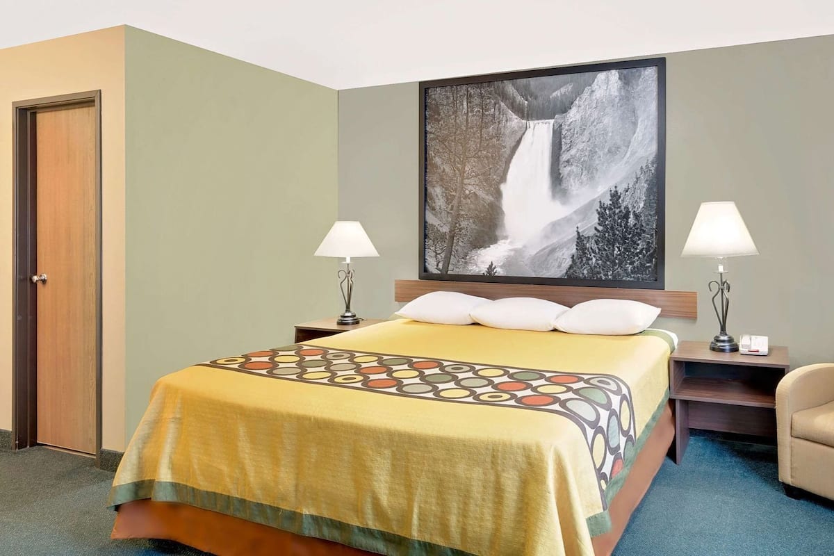 A hotel room with a bed, two bedside lamps, a chair, a large wall-mounted picture of a waterfall, and a door.