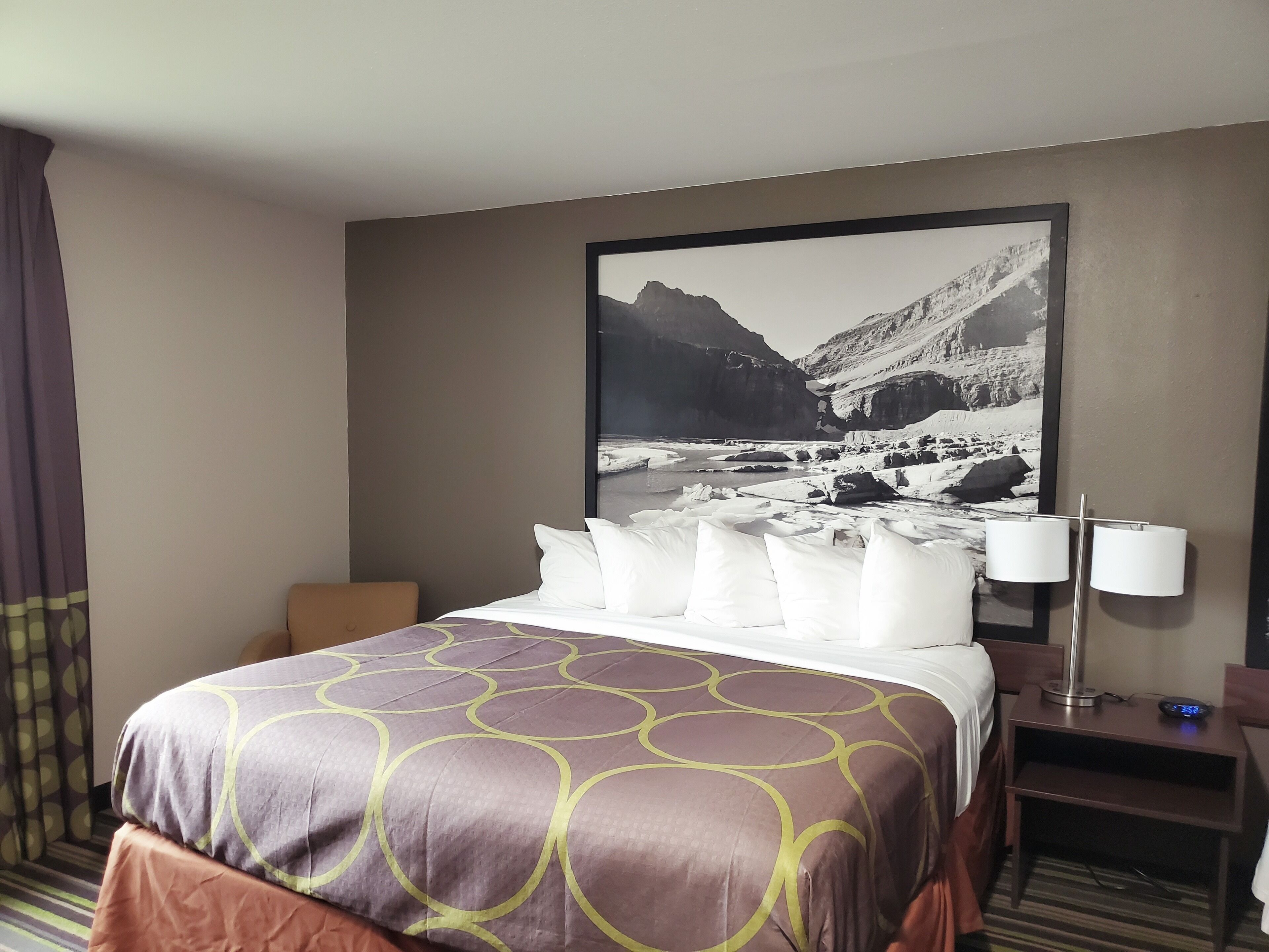 Ein Hotelzimmer mit einem Bett, einem Nachttisch mit Lampe und einem großen gerahmten Bild einer Berglandschaft.