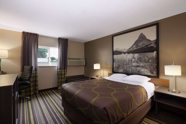 A hotel room with a king-sized bed, a desk with a chair, a large window with curtains, a wall-mounted air conditioner, and a framed picture of a mountain landscape.