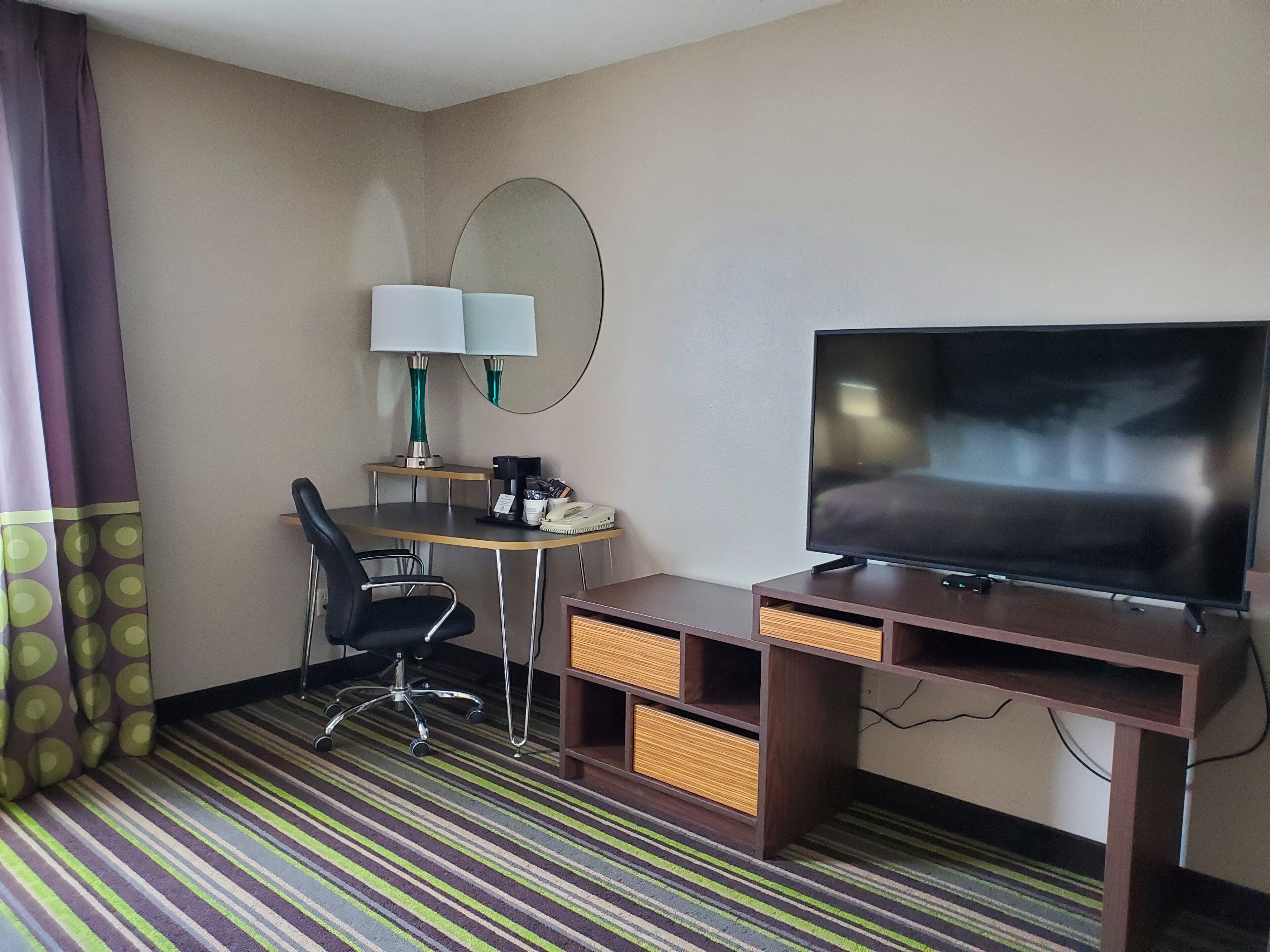 A hotel room with a bed, a TV, a desk with a lamp, a chair, and a striped carpet.