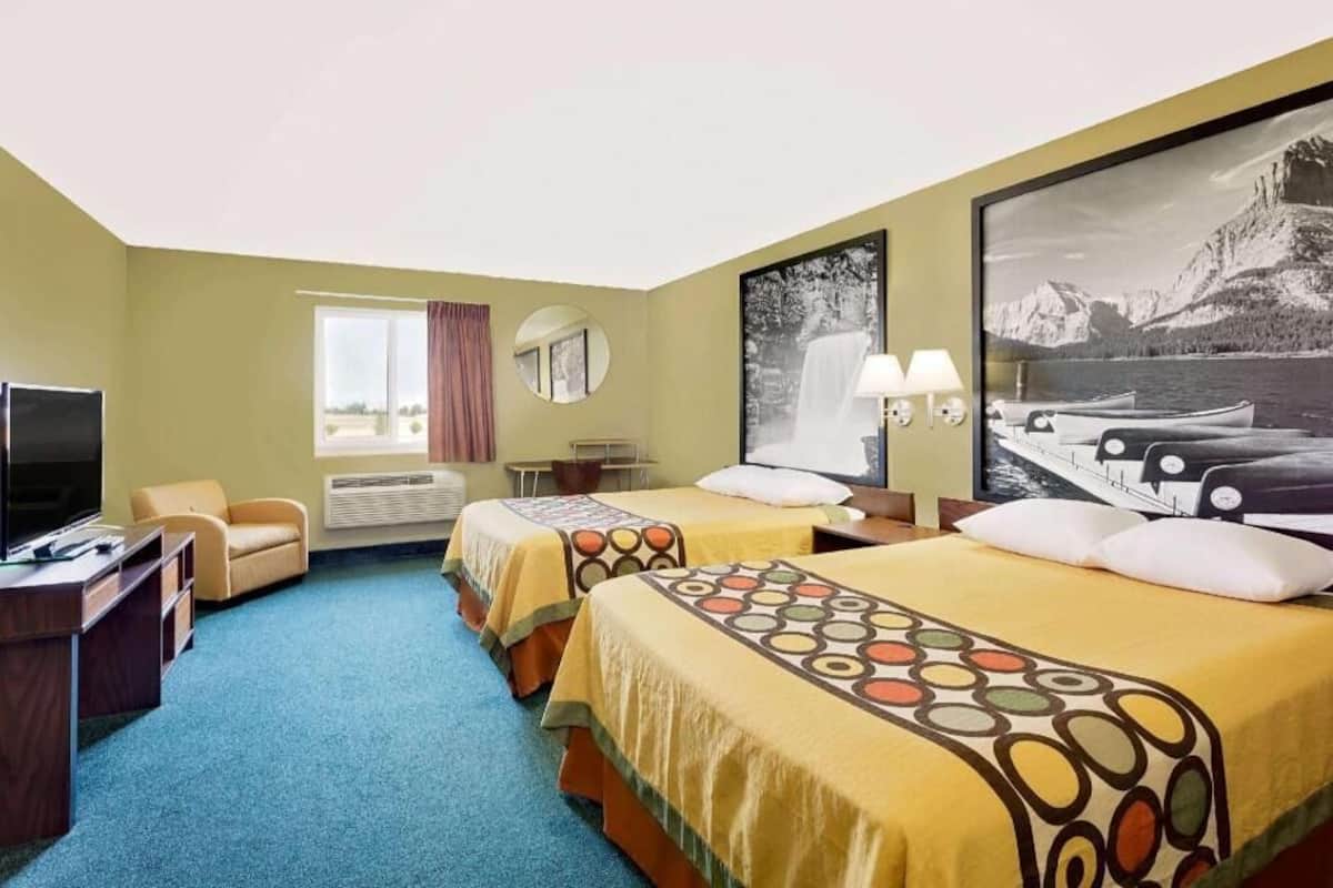 Ett hotellrum med två sängar, ett skrivbord, en tv, ett fönster med gardiner och en stor, inramad tavla på väggen.