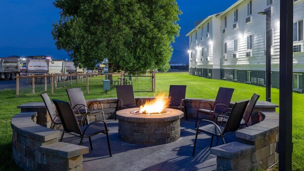 An outdoor fire pit with chairs, surrounded by stone walls, in a well-lit area with a building in the background.