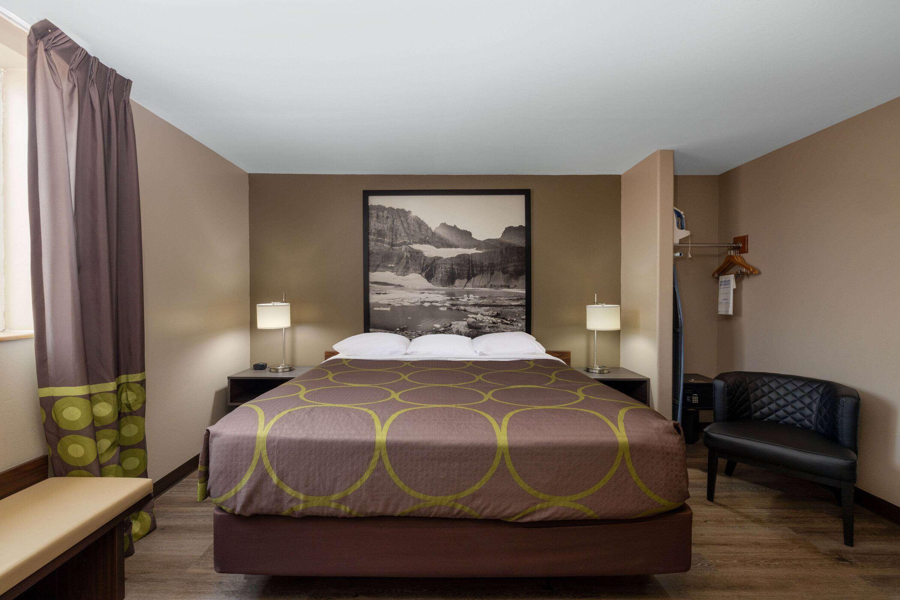Habitación de hotel con cama, dos mesitas de noche, una silla, escritorio, lámpara, un cuadro en la pared y ventana con cortinas.