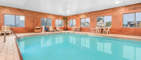 A swimming pool with wooden walls and large windows, surrounded by lounge chairs and a small table.