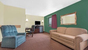 Une chambre d’hôtel avec un fauteuil bleu, un canapé beige, un bureau avec un ordinateur, une télévision et un miroir au mur.