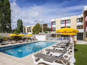 A swimming pool with sun loungers and yellow umbrellas, surrounded by a building with multiple windows and a white facade. - ibis Styles Nice Cap 3000 Airport (Saint-Laurent-du-Var)
