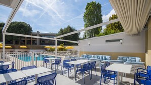 A poolside area with blue chairs and tables, a pool, and a covered seating area with yellow umbrellas. - ibis Styles Nice Cap 3000 Airport (Saint-Laurent-du-Var)