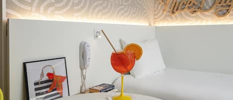 Siististi järjestetty hotellihuone, jossa on sänky, yöpöytä, jossa on juoma, puhelin ja kehystetty kuva.