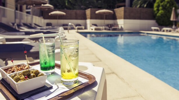 A poolside setting with a tray of drinks and olives, a lounge area with umbrellas, and a clear view of the pool. - ibis Styles Nice Cap 3000 Airport (Saint-Laurent-du-Var)