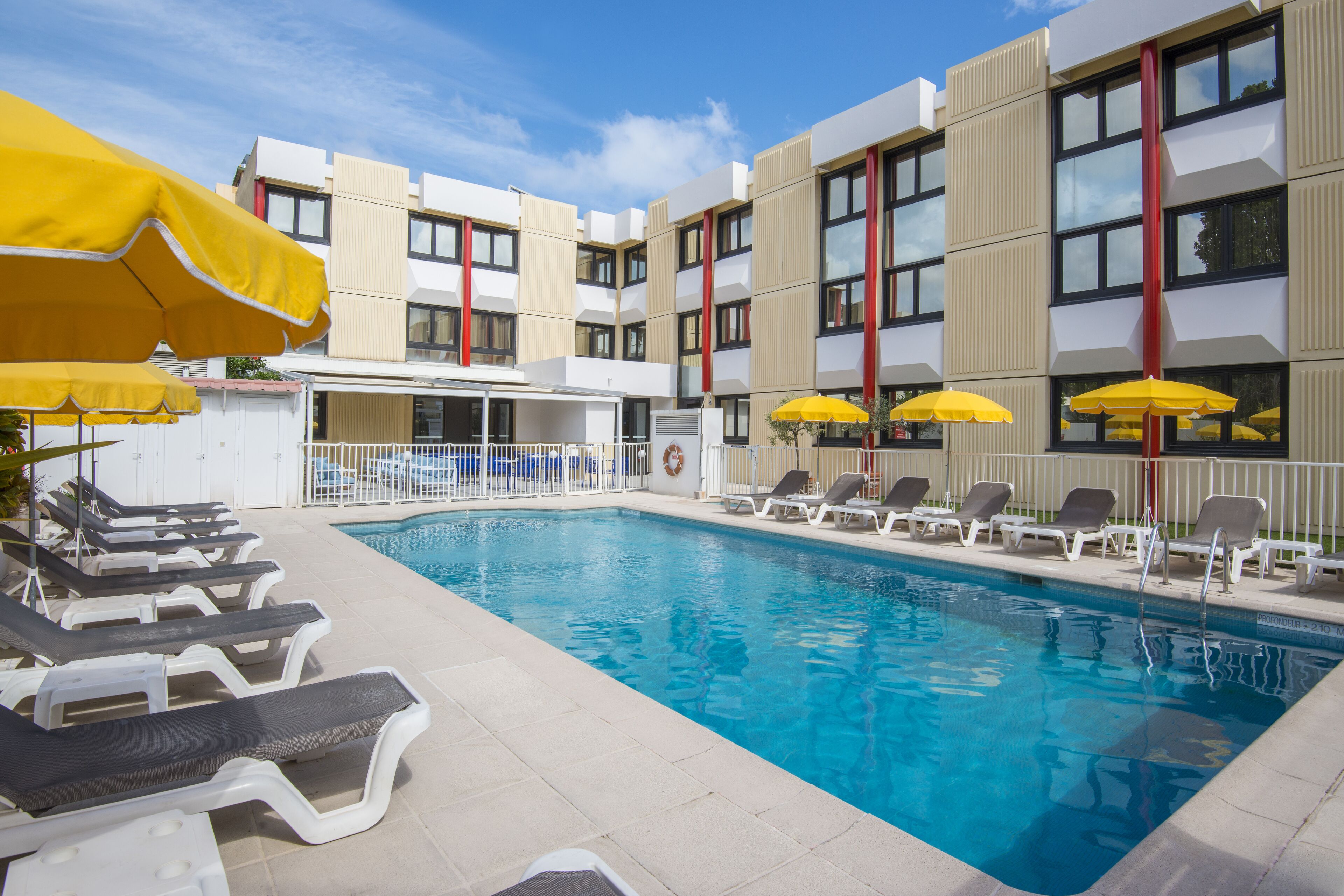A swimming pool with sun loungers and yellow umbrellas, surrounded by a modern building with balconies.