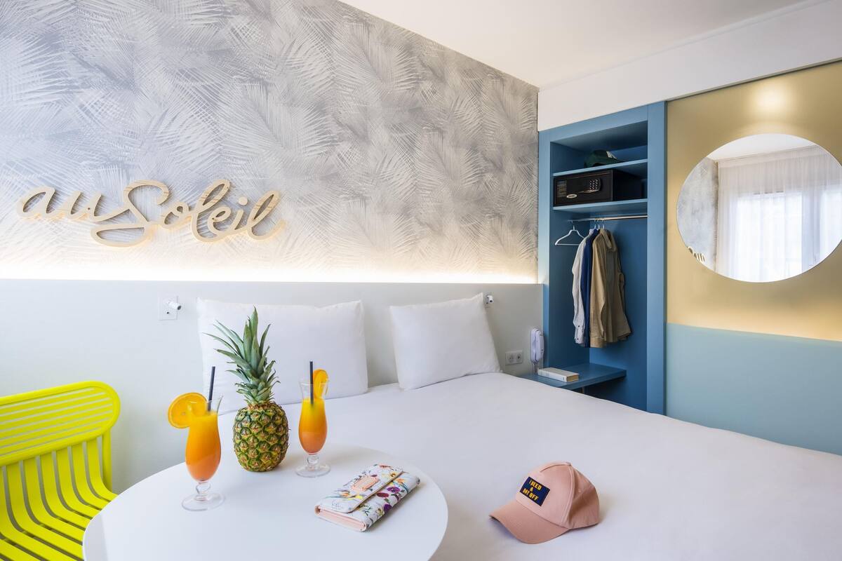 A hotel room with a bed, a round table with drinks and a pineapple, a bright yellow chair, and a wardrobe with clothes.