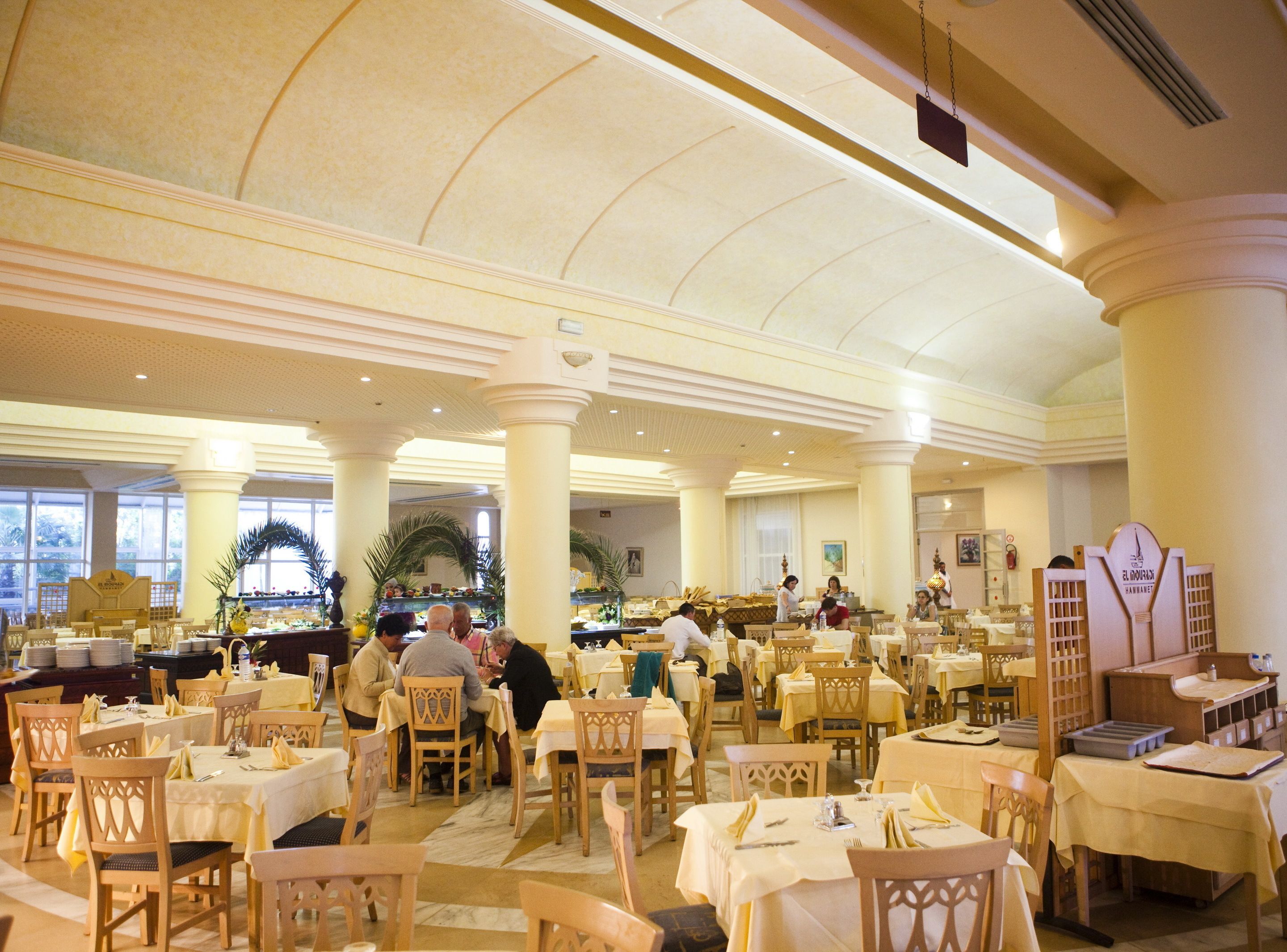 A spacious dining area with multiple tables and chairs, large windows, and a high ceiling with decorative moldings.