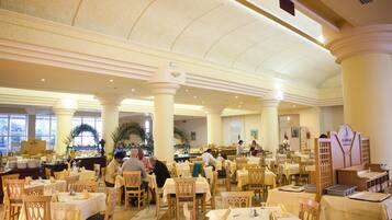 A spacious dining area with multiple tables and chairs, large windows, and a high ceiling with decorative moldings.