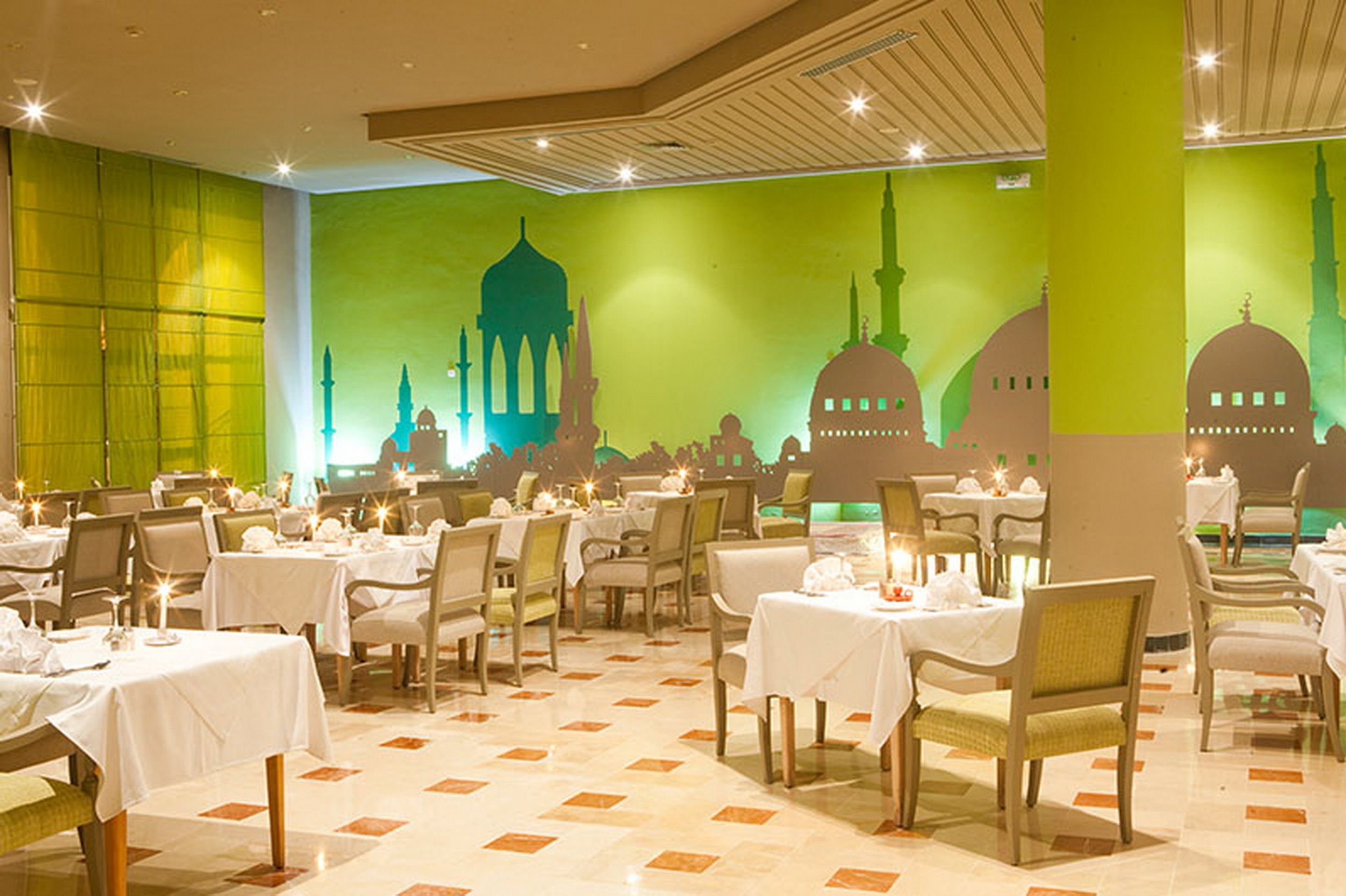 A restaurant with tables set for dining, a green wall with mosque silhouettes, and a tiled floor.