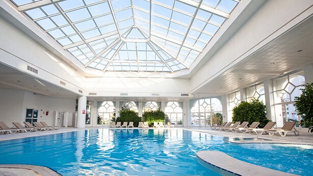Une grande piscine intérieure dotée d’un toit en verre, entourée de chaises longues et de plantes en pot.