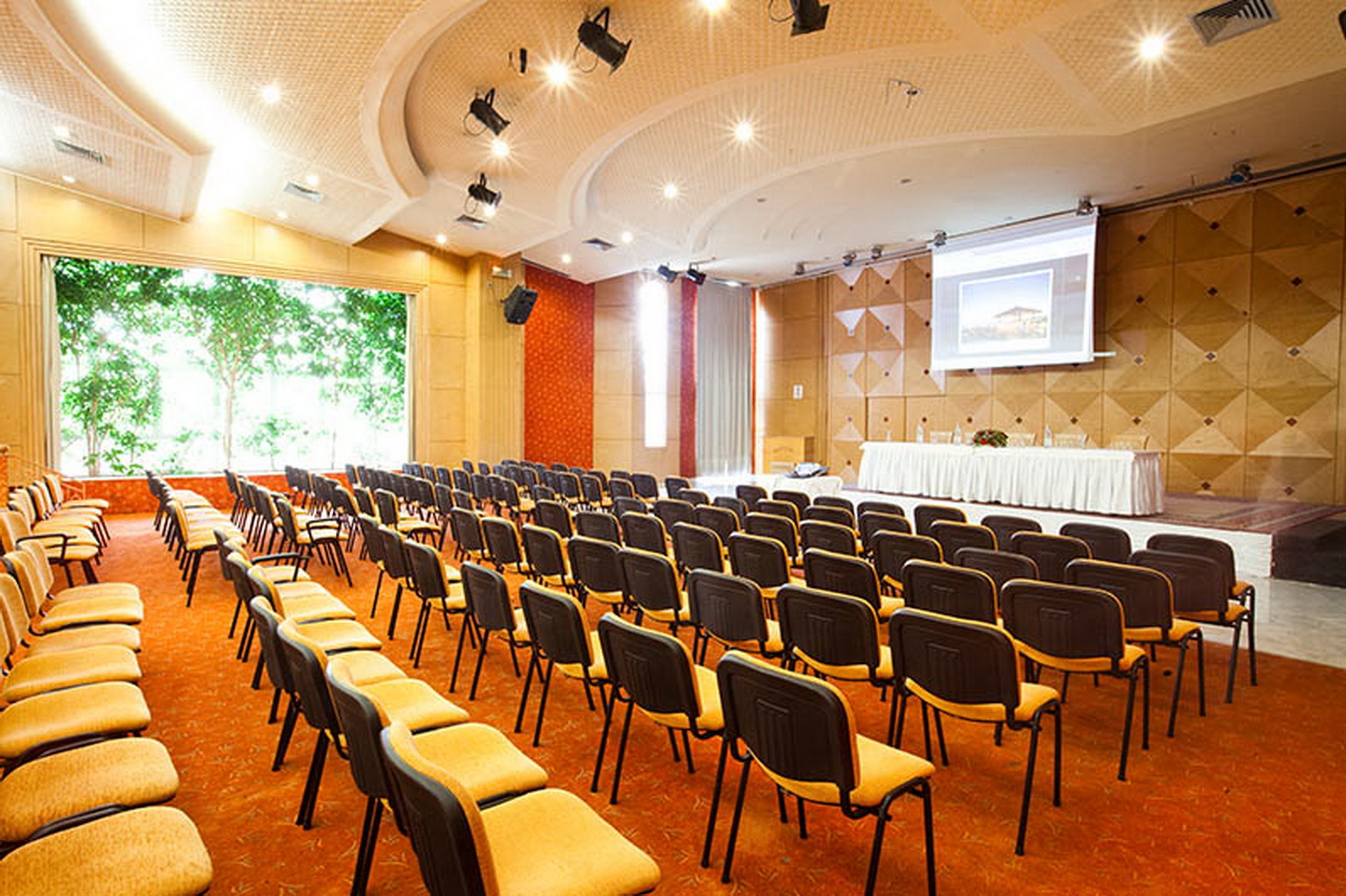 Une salle de conférence avec des rangées de chaises, une scène avec une table et un écran de projection.