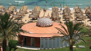 Une plage avec des parasols en chaume, un palmier et un bâtiment surmonté d’un dôme.
