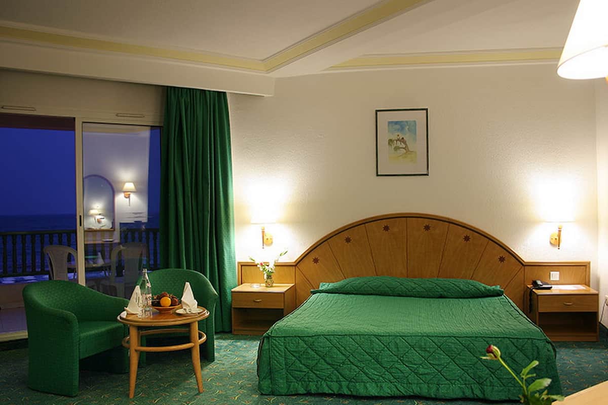 Habitación de hotel con cama, una silla, una mesa con botella y vasos, una mesita de noche con lámpara, y vista al océano a través de una ventana.