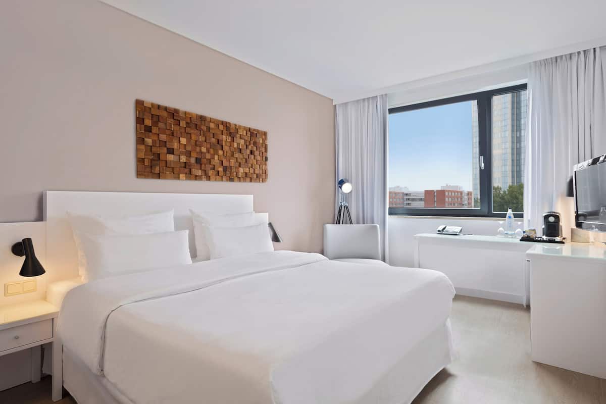 Une chambre d’hôtel avec un grand lit, un bureau, une lampe, une machine à café et une vue sur la ville, visible à travers une grande fenêtre.