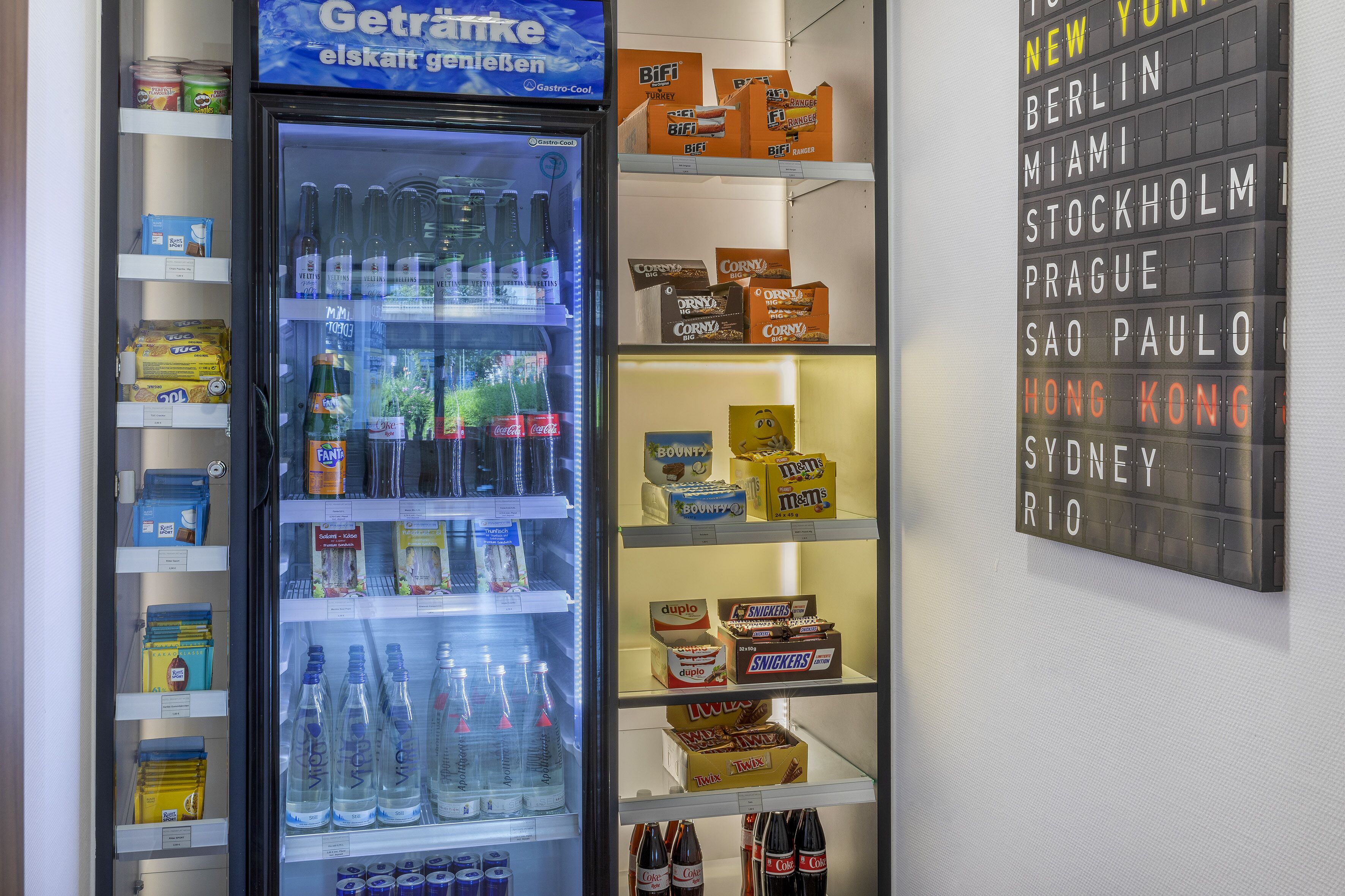 Ein Getränkeautomat mit verschiedenen Getränken und Snacks, umgeben von Regalen voller verpackter Lebensmittel.