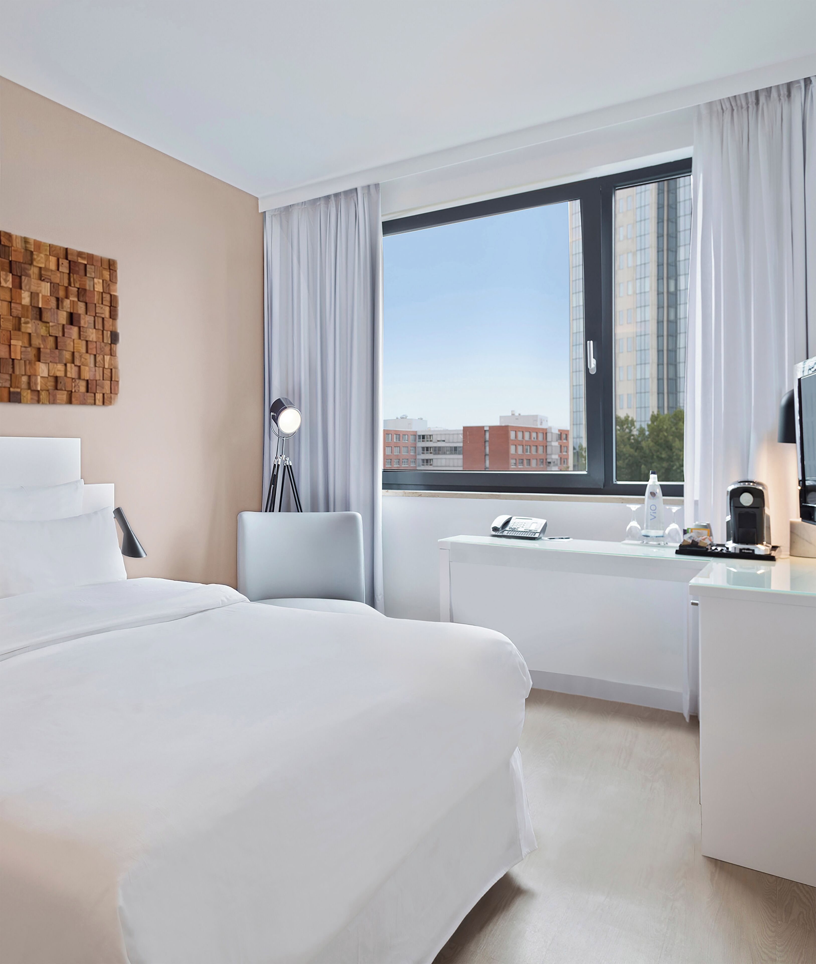 Ein Hotelzimmer mit Bett, Schreibtisch, Stuhl, Lampe, einem Fenster mit Vorhängen und Blick auf die Stadt.