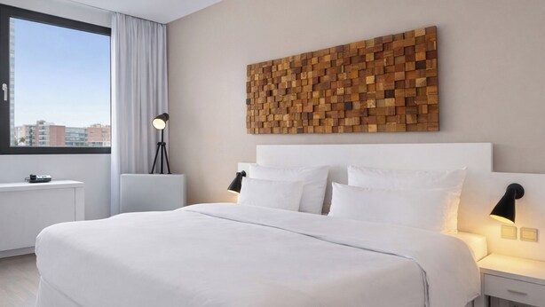 A hotel room with a large bed, a city view, a wooden wall art piece, and bedside lamps. - Hotel Frankfurt Messe affiliated by Meliá (Frankfurt)