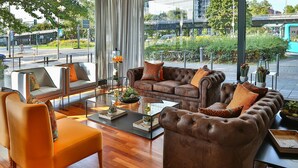 A modern living room with a sofa, armchair, and coffee table, featuring a view of a city street and greenery outside. - Hotel Frankfurt Messe affiliated by Meliá (Frankfurt)
