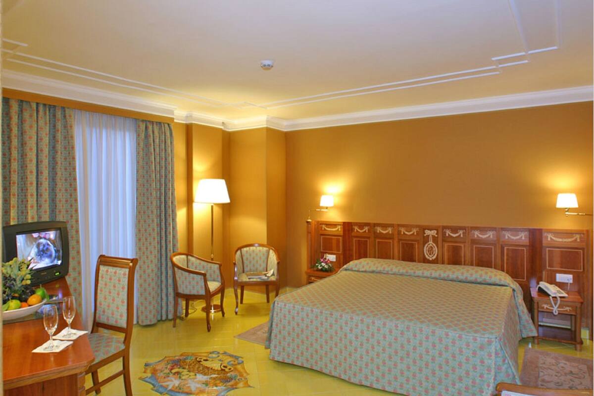 A hotel room with a bed, two chairs, a desk with a television, and a nightstand with a lamp.