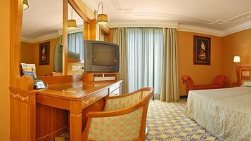 Una habitación de hotel con un escritorio de madera, televisión, cama, silla y un ventanal grande con cortinas.