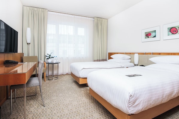 A hotel room with two beds, a desk, a chair, a small table with a plant, and a TV.