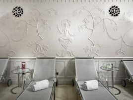 Um espaço de spa com duas poltronas reclináveis, uma mesinha com um copo, e uma parede decorativa com motivos florais.
