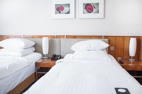 A hotel room with two single beds, a wooden headboard, bedside tables with lamps, a phone, and framed artwork on the wall.