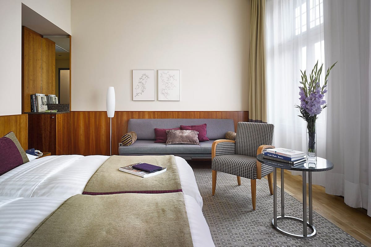 A hotel room with a bed, a sofa, a chair, a small table with books, a vase with flowers, and two framed pictures on the wall.