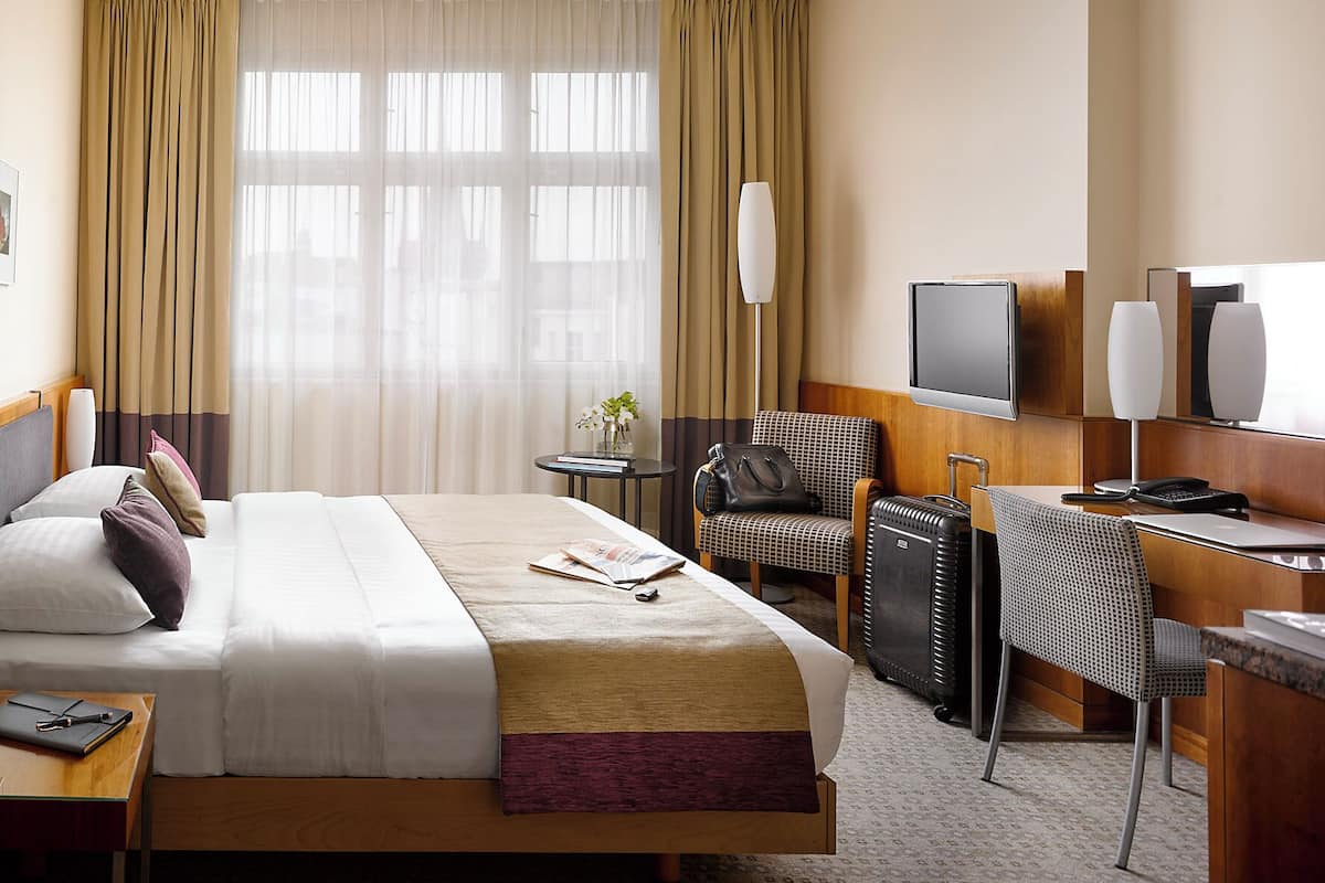 A hotel room with a bed, a desk with a chair, a TV, and a window with curtains.