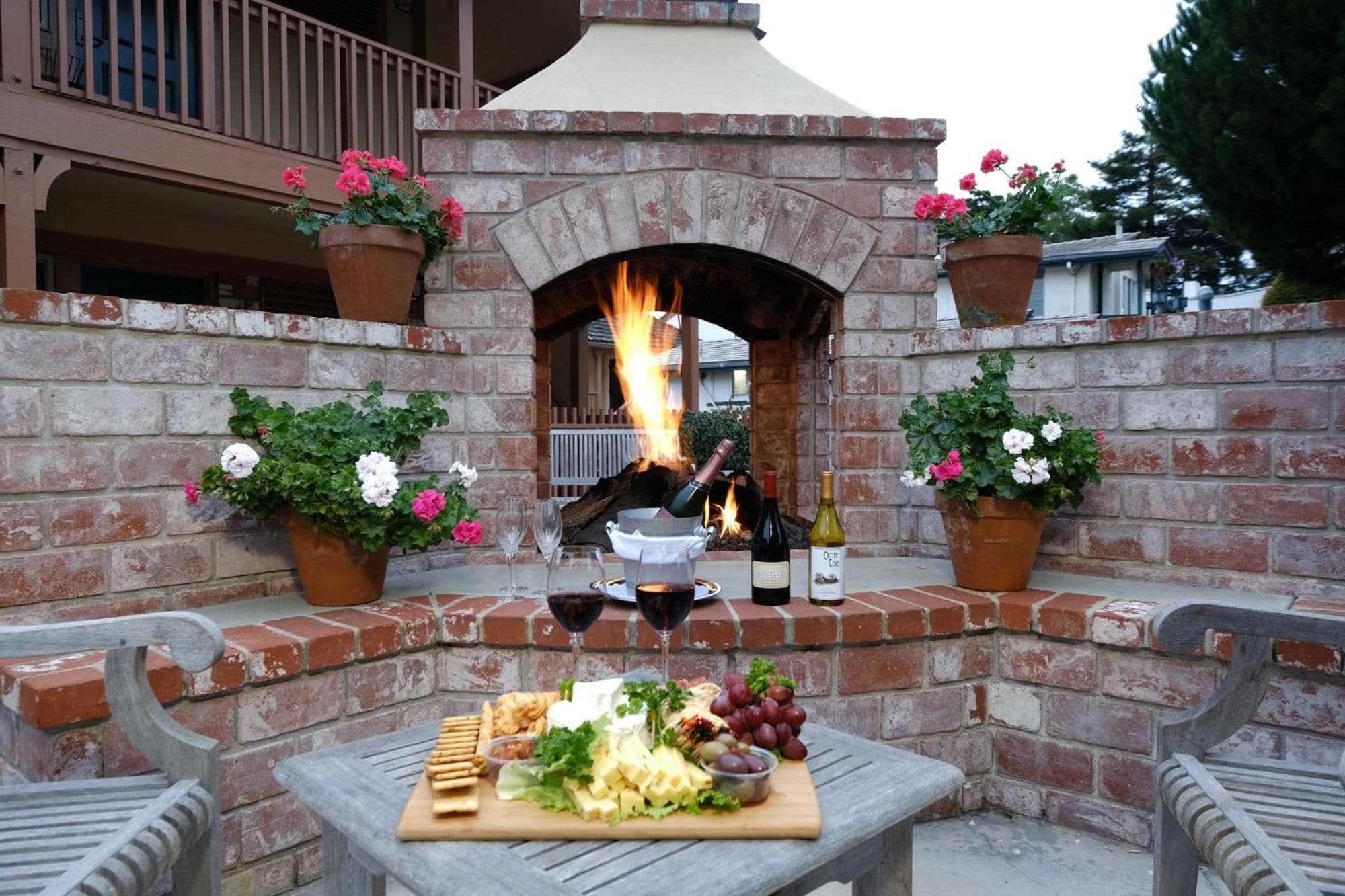 An outdoor brick oven with a fire, surrounded by potted plants and a table with food and drinks.