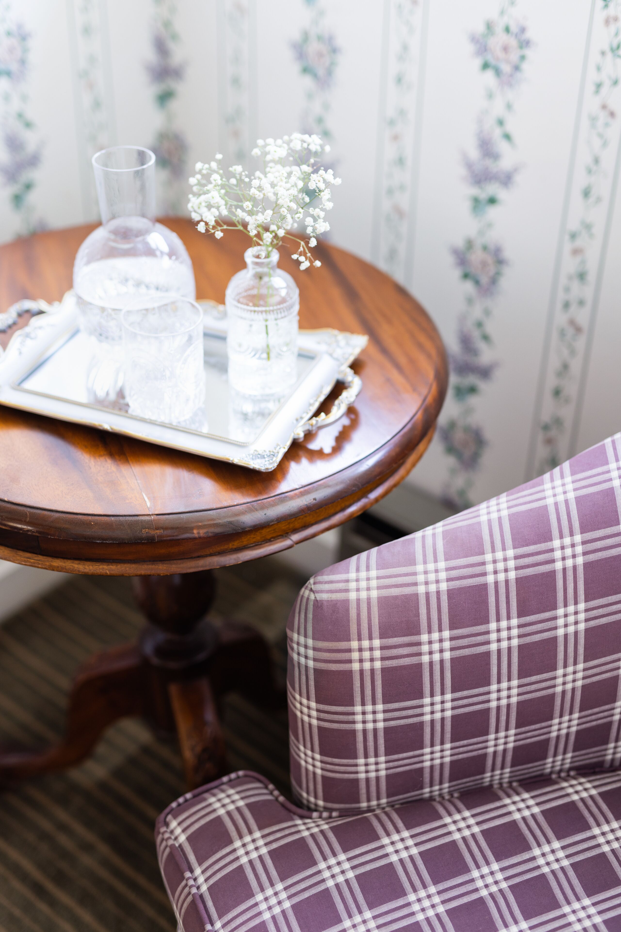 A round wooden table with a tray holding a glass of water, a glass of wine, and a small vase with flowers. A plaid chair is partially visible in the foreground.