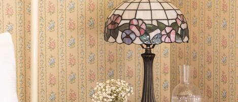 A bedroom with a floral wallpaper, a bedside table with a lamp, a vase with flowers, and a nightstand with a glass bottle.