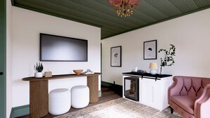 Una sala moderna con una consola de madera, un sillón rosa, un televisor de pantalla plana grande e impresiones botánicas enmarcadas en la pared.