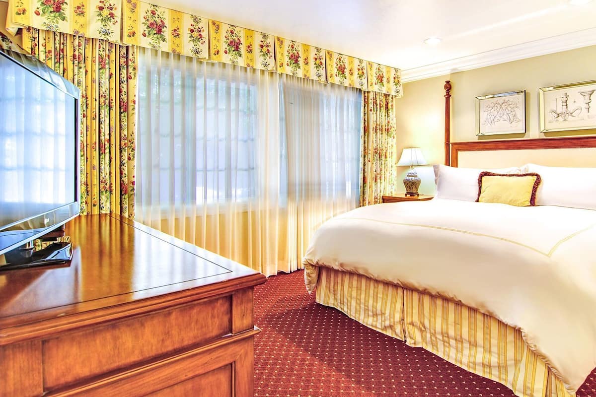 Quarto de hotel com uma cama grande, uma televisão sobre uma escrivaninha de madeira e uma janela com cortinas florais.