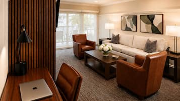 A hotel room with a wooden desk, a laptop, a sofa, two armchairs, a coffee table with a vase of flowers, and two wall-mounted lamps.