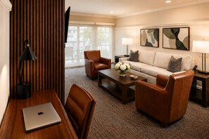 A hotel room with a wooden desk, a laptop, a sofa, two armchairs, a coffee table with a vase of flowers, and two wall-mounted lamps. - Wayside Inn (Carmel)