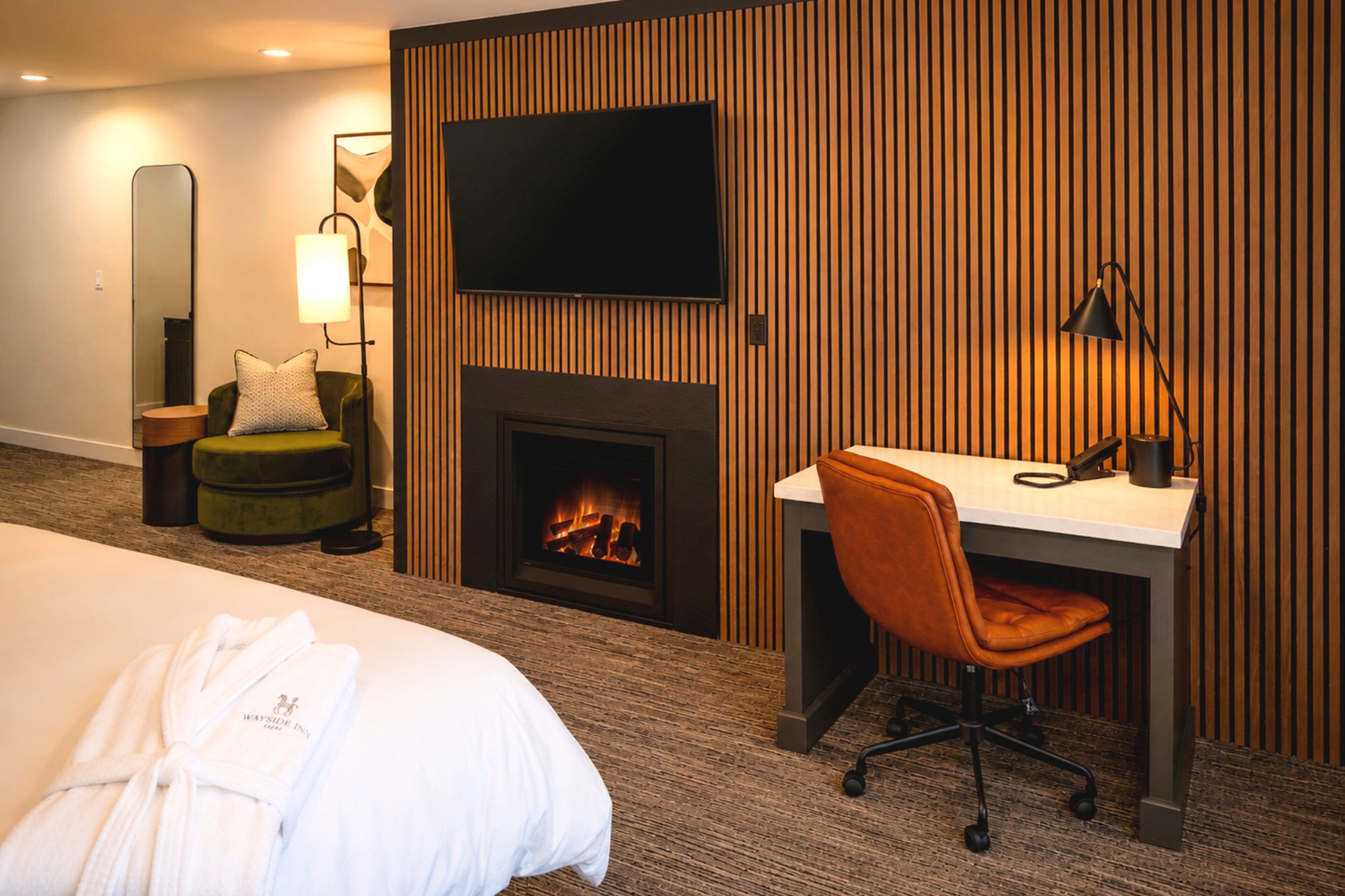 A hotel room with a bed, a desk, a chair, a television, a fireplace, a lamp, and a mirror.