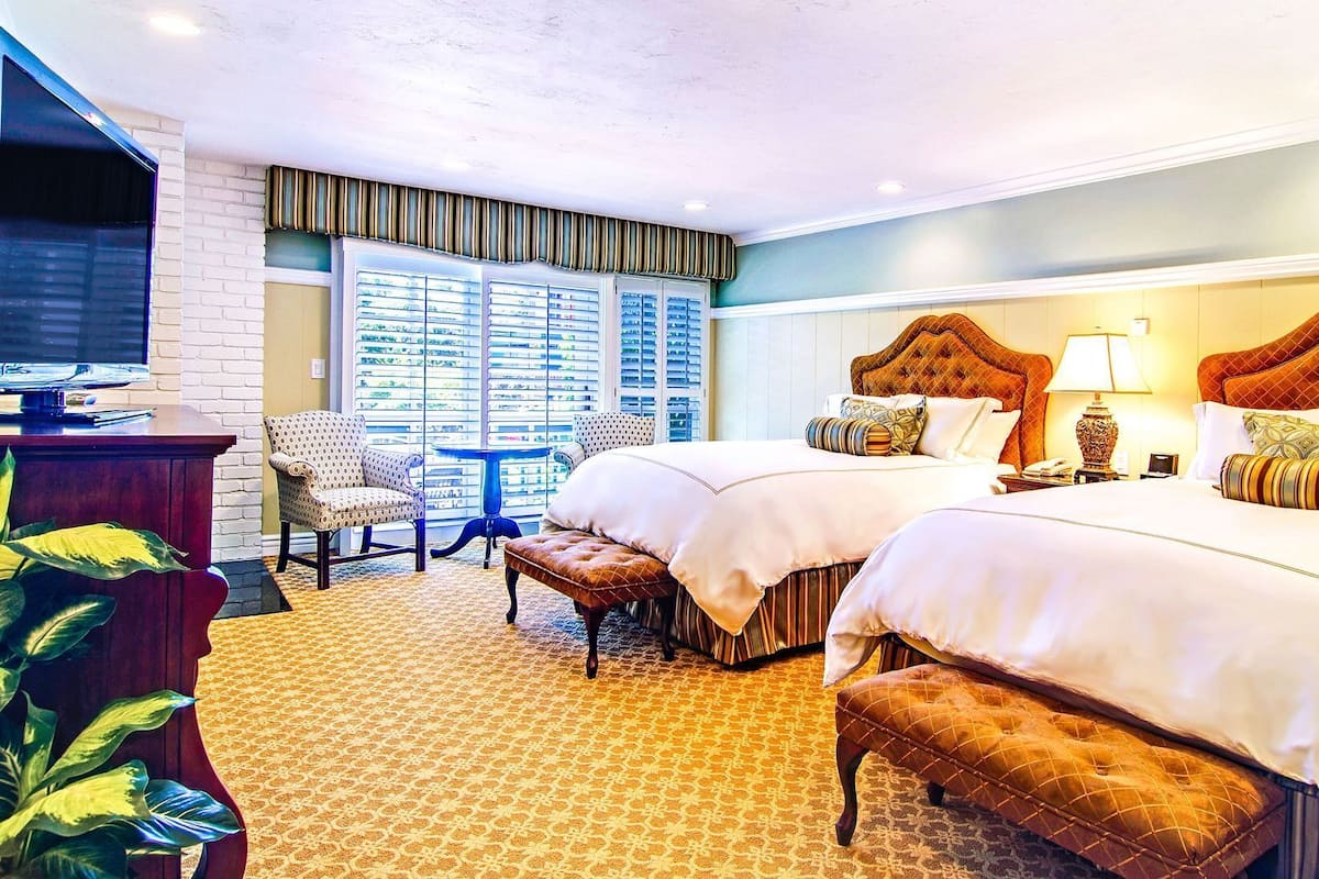 Quarto de hotel com duas camas, uma televisão, uma escrivaninha e uma janela com persianas.