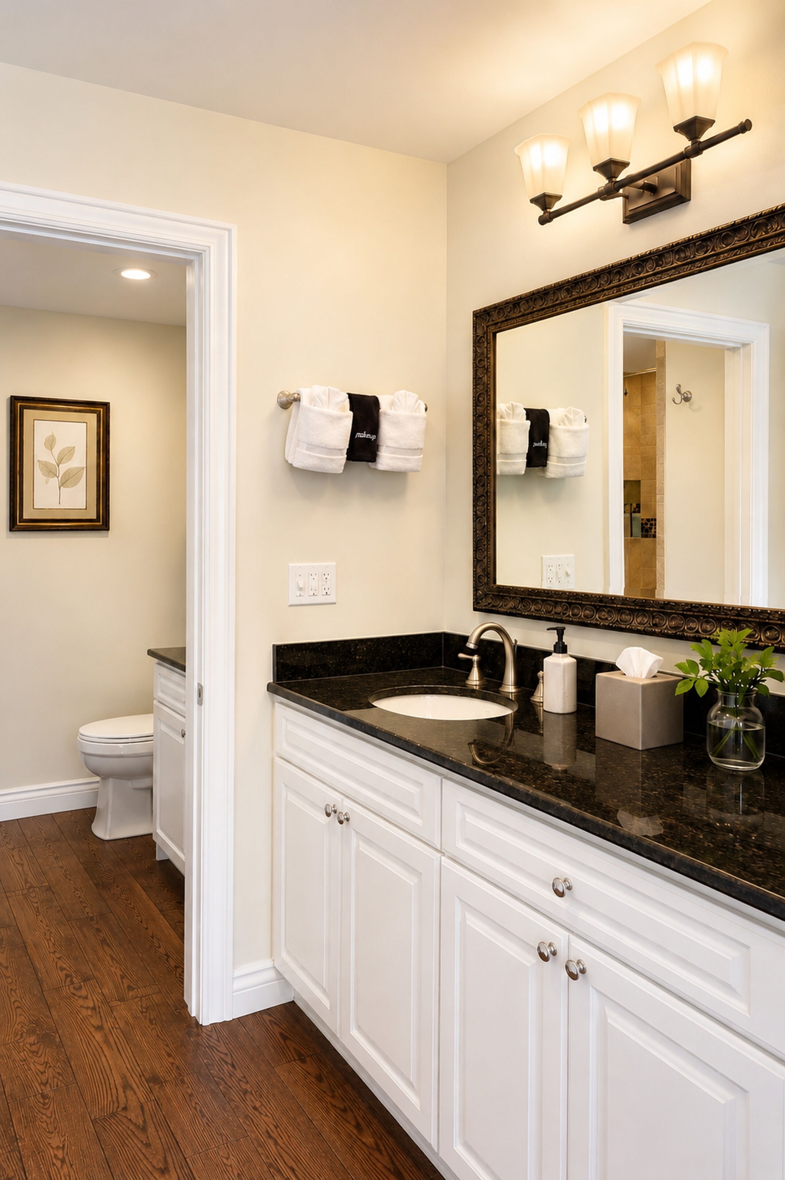 A bathroom with a white vanity, a black countertop, a large mirror with lights, a toilet, and a framed picture on the wall.