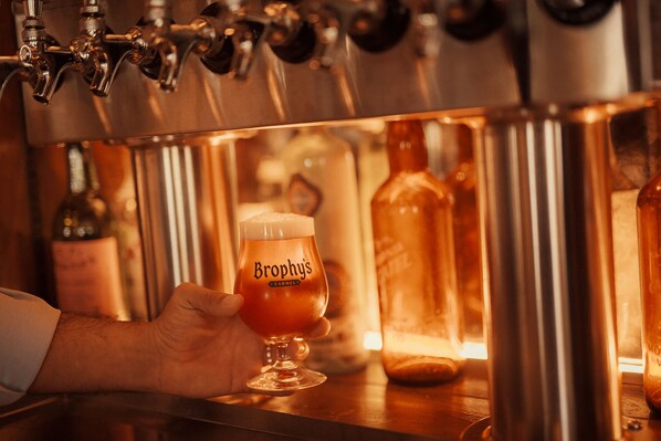 A hand holding a glass of beer with the label "Brophy's" in a bar setting.