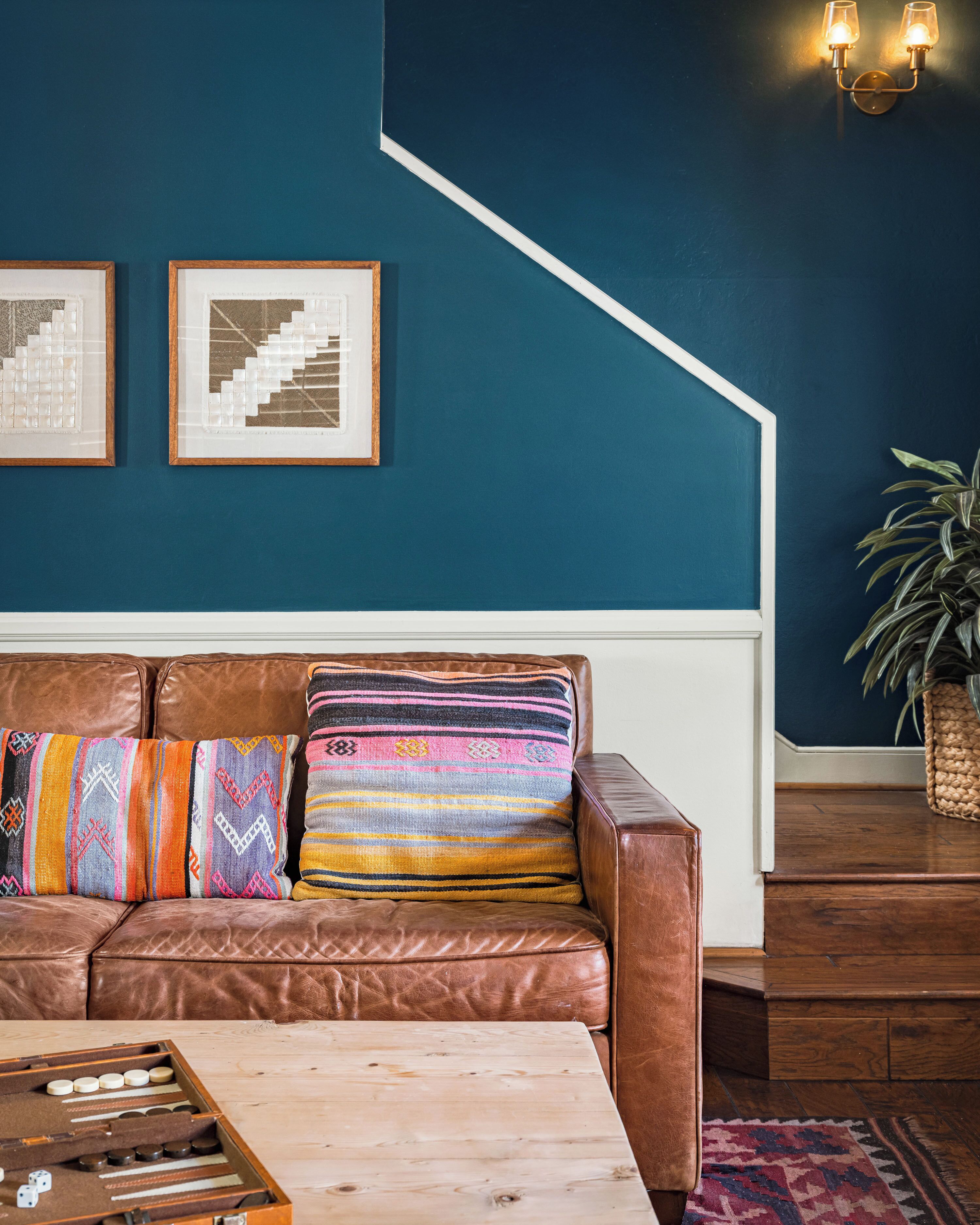 A brown leather sofa with colorful pillows, a wooden coffee table, and a framed abstract painting on a teal wall.