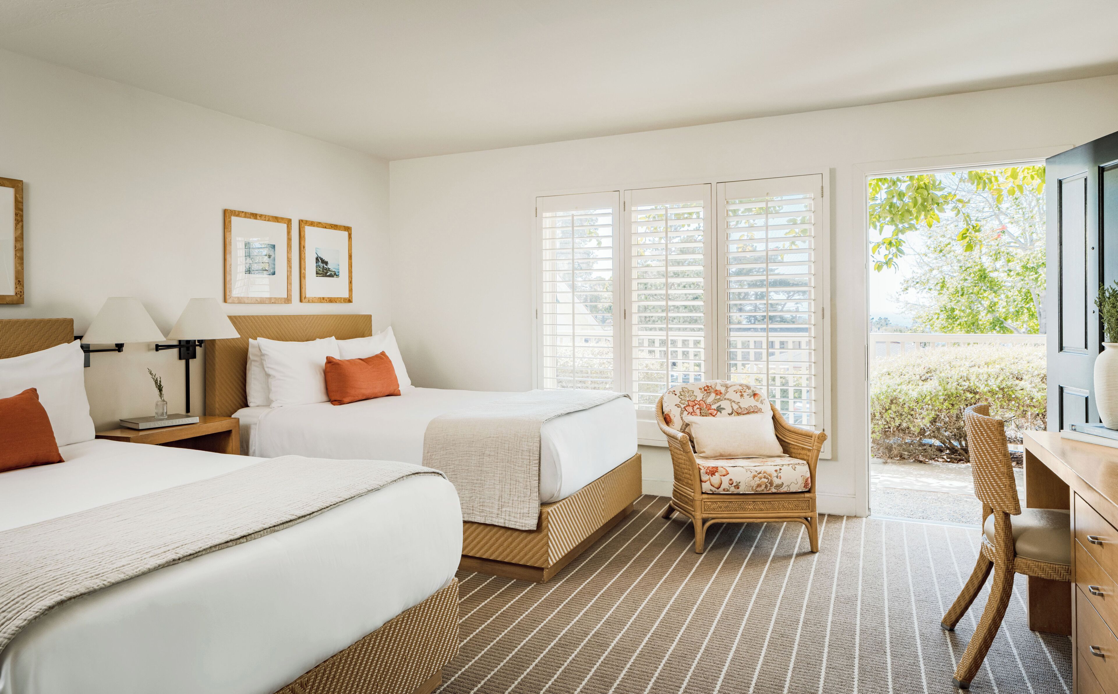 A hotel room with two beds, a wicker chair, a desk, and a view of greenery.